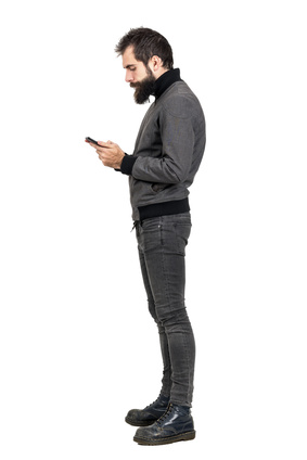 Side view of stylish bearded hipster typing on his smartphone. Full body length portrait isolated over white studio background.